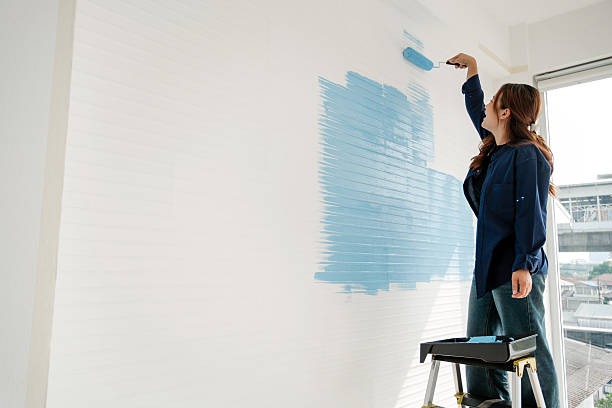 istock A person on a step stool paints a white wall light blue with a roller in a bright room near a large window, suggesting a home DIY renovation project. 2246311327