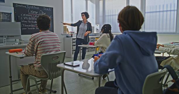 istock Female Teacher Stands Near Blackboard Explaining Coding Assignment to Group of Students in Bright Classroom 2246313165