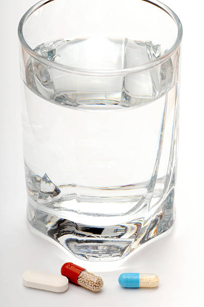 Vertical photo of a clear, glass of water with three different colorful pills and capsules placed in front of it  daily healthcare routine.