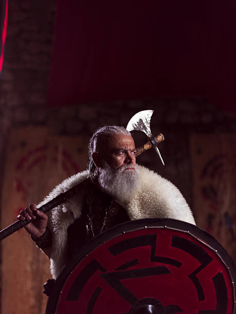 istock A resolute Viking chieftain, with a white beard and fur cloak, stands holding a formidable axe over his shoulder and a red and black round shield, embodying strength and ancient resolve. 2246321184