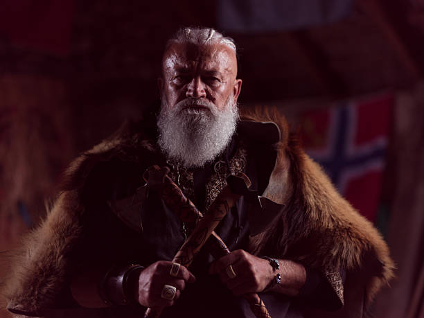 istock A formidable Viking chieftain with a striking white beard and braided hair, draped in a rich fur cloak, holds two crossed axes, gazing intensely into the camera in a dimly lit, rustic setting. 2246321256