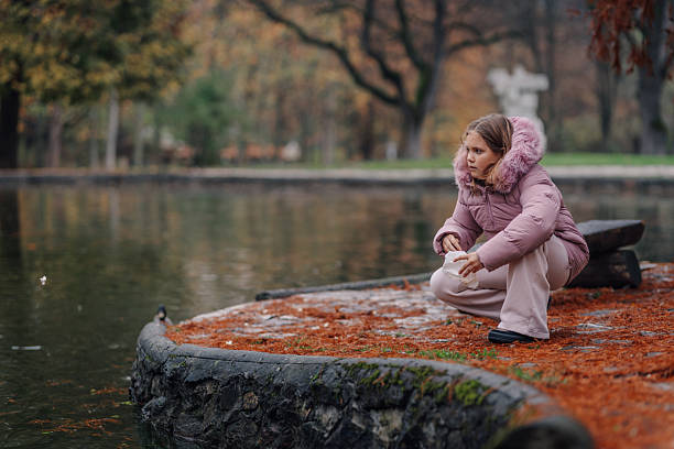 Young girl wearing a warm winter jacket crouching by a pond, feeding birds with attention during a cold autumn day