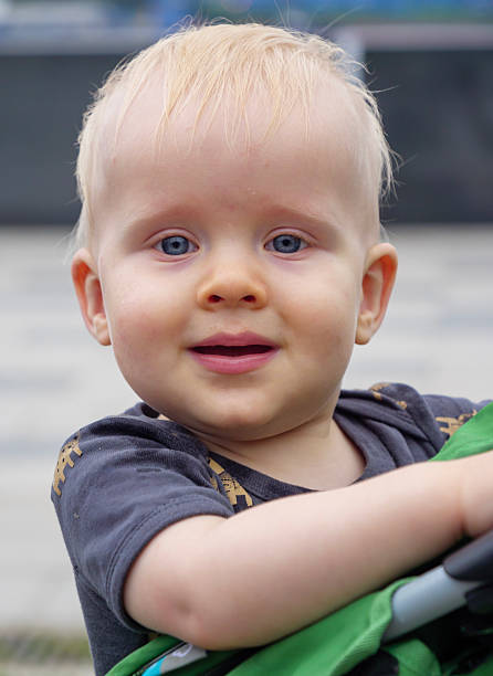 A cheerful toddler with light blond hair and blue eyes gazes curiously while nestled comfortably in a stroller on a warm day outside.