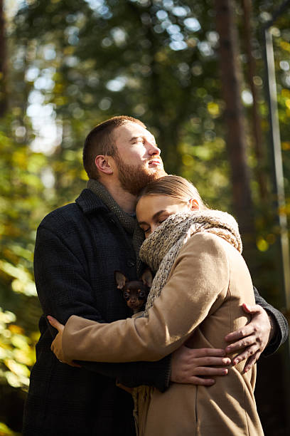 Caucasian young adult man embracing Caucasian young adult woman outdoors in forest, woman resting head on man's chest while holding small dog, both appearing relaxed and affectionate