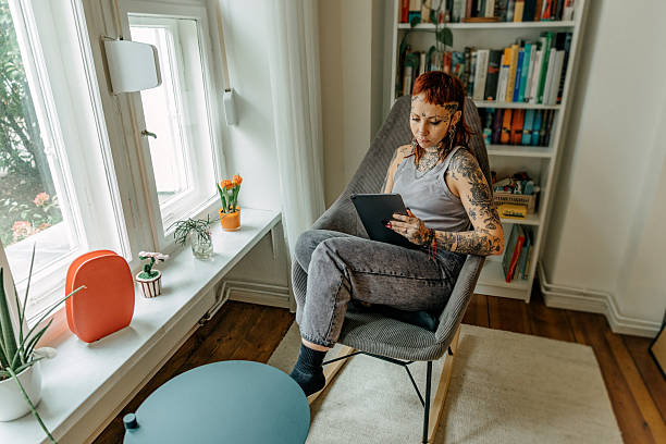 istock Tattooed woman using digital tablet while sitting in armchair near window 2246338374