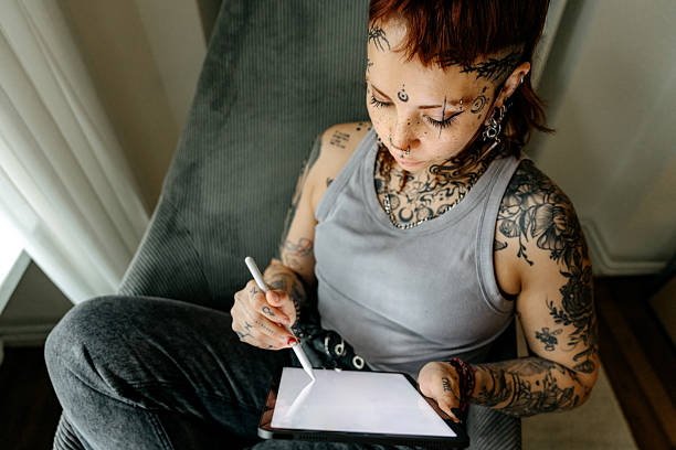 Stylish young woman with tattoos sitting comfortably in an armchair, focused on her digital tablet, showcasing modern technology use, creativity, and a relaxed home environment