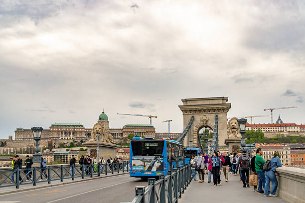 Budapest, Hungary: A diverse group of pedestrians, including men, women, and children of various ages, walks across the historic Chain Bridge, known as Széchenyi lánchid. The bridge features ornate neoclassical architecture with prominent lion statues at its ends and suspension cables arching overhead. In the background, construction cranes indicate ongoing renovation work near the riverbank. The scene shows people on the pedestrian walkway in casual summer attire carrying bags and pushing pushchairs.