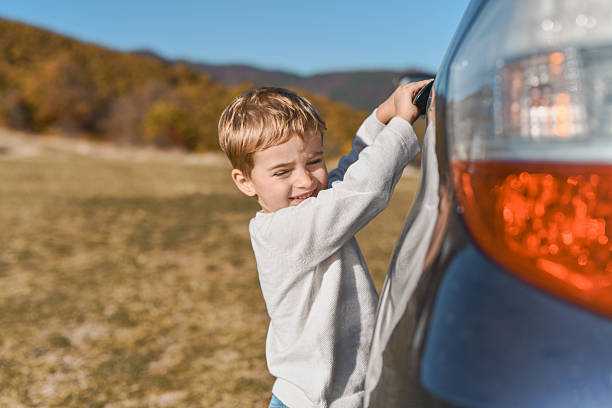 A cheerful boy smiles as he pulls on a car door in a sunny, open field. Golden hills and clear blue sky create a warm, outdoor family moment.