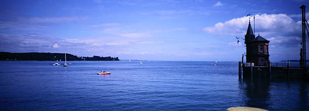 Tranquil summer on Lake Constance (Bodensee) in Konstanz, Germany. A historic harbor tower with a pointed roof stands sentinel over calm deep-blue waters dotted with sailboats and a small motorboat carrying two passengers. The distant tree-lined shoreline fades into the horizon under a vast sky with scattered clouds, capturing the peaceful maritime charm and ambiance of this iconic European lake.