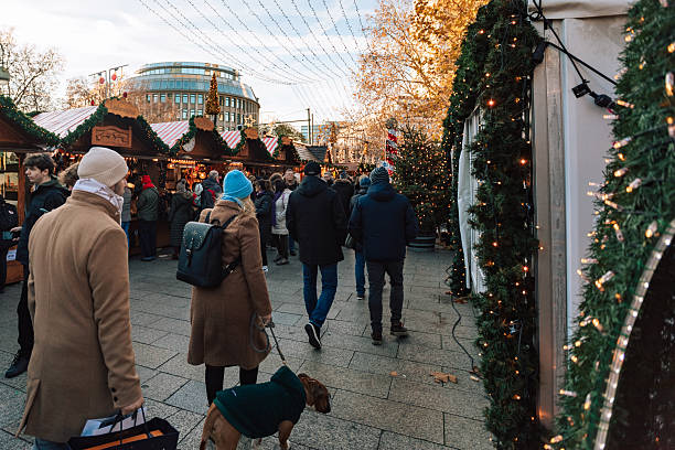 Berlin, Germany – November 30, 2024: People with winter coats and a dog walk among the decorated stalls at the Christmas market on Breitscheidplatz in Berlin. Warm lights and garlands create a festive city atmosphere.