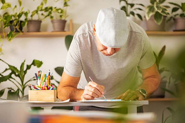 A thoughtful and focused senior man is intensely drawing at his desk, captured in a close-up portrait surrounded by various lush houseplants in his bright home studio.