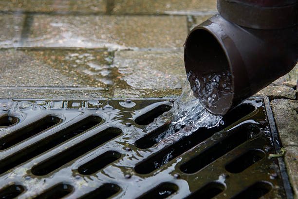 Close-up of a brown drainage pipe releasing water into a metal grate in a courtyard. Concept of rainwater management, plumbing, and urban infrastructure.
