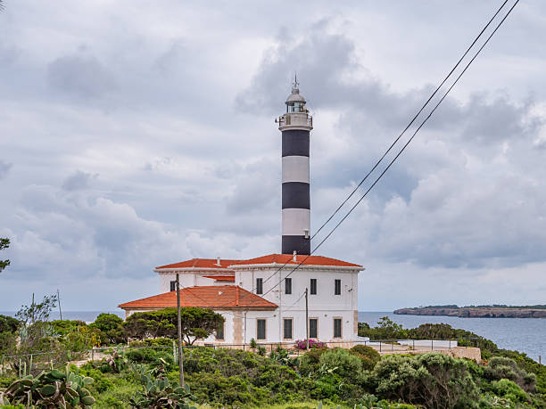 istock Portocolom lighthouse, Mallorca, lighthouse stands on a coastal island, surrounded by lush greenery and a serene sea under a cloudy sky, Majorca 2246382296