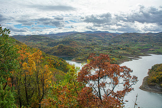 istock Colorful Autumn Trees Overlooking a Winding Lake Amid Hills, Green Fields, and Distant Mountains Under a Cloudy Sky 2246384348