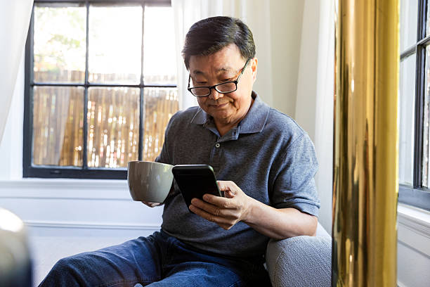 istock Elderly Asian man using his smartphone and holding a cup of coffee while relaxing indoors. 2246399196