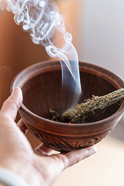 Hand holds a burning mugwort smudge stick in a clay bowl, producing purifying herbal smoke.