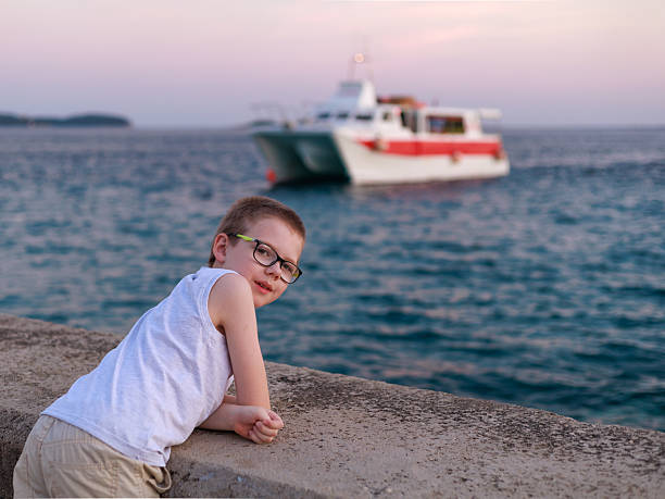 Boy in glasses leaning on seaside wall looking at boats during family trip, soft sunset light, copy space. Concept of childhood curiosity, travel memories, summer relaxation.