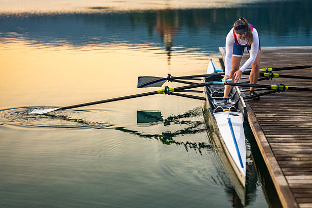 An american shot photo of a female rower in a double scull prepares for a training session on a serene lake at sunset. The calm water reflects the golden hues, emphasizing focus and teamwork in sports training.