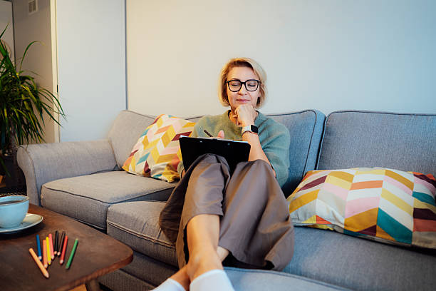 Middle aged woman planning goals for next new year while sitting on the sofa at home. Self-reflection, plans, action, checklist concept. Personal Year review. Coaching tools. Finding Balance in Life