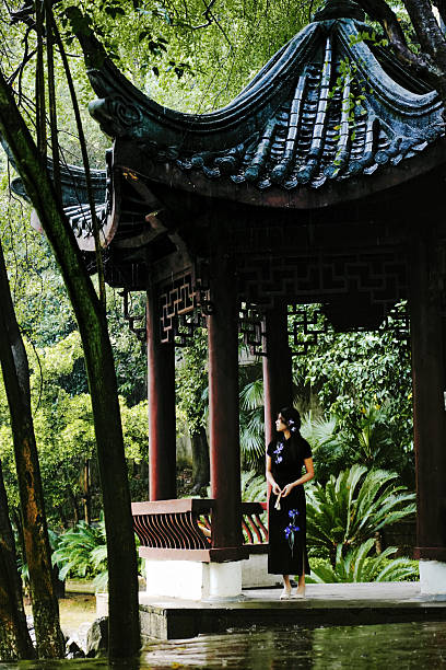 She stands beneath the curved eaves of an ancient pavilion, surrounded by lush vegetation. The damp, misty air of the rainy day lingers softly in the scene.
