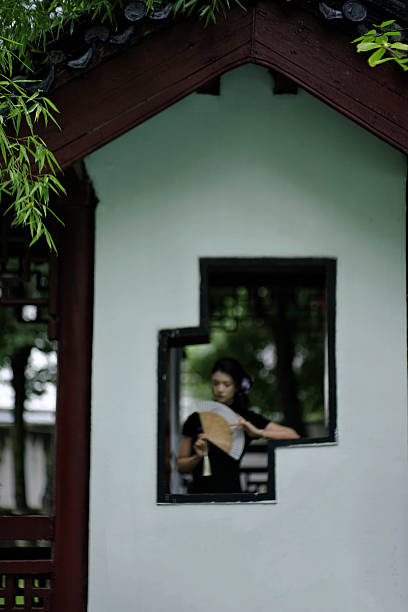 Peeking through the hollowed lattice window of the garden, she gently waves a folding fan. The sense of negative space in the traditional architecture enhances the subtle charm of the moment.