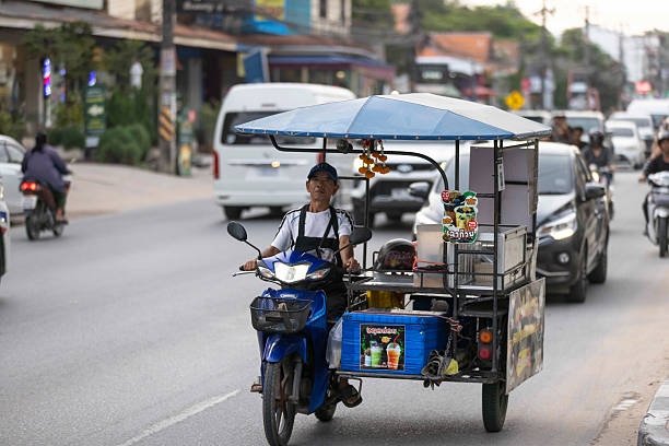 11 November 2025, Ko Samui - Thailand.A typical traffic scene in Koh Samui shows a car, a motorcycle, and a tricycle-style food stall sharing the same road. The mix of regular vehicles and mobile street vendors moving side by side is a common and familiar sight in Thailand, reflecting the country’s everyday street culture.