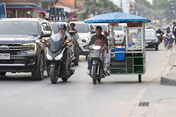 11 November 2025, Ko Samui - Thailand.A typical traffic scene in Koh Samui shows a car, a motorcycle, and a tricycle-style food stall sharing the same road. The mix of regular vehicles and mobile street vendors moving side by side is a common and familiar sight in Thailand, reflecting the country’s everyday street culture.