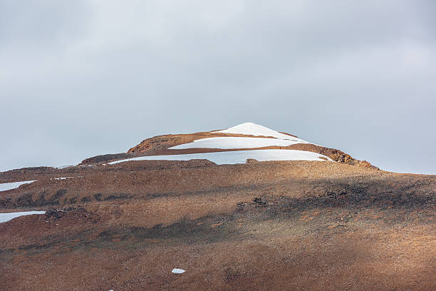 istock Sunlit snowy pointy peak under gray cloudy sky. Triangle shaped snow mountain top in bright sun. Stony hill of pyramid shape in changeable weather. Sharp rocky snowbound pinnacle in overcast sunny day 2246456065
