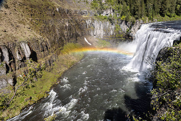 A dramatic view of Upper Mesa Falls in Idaho, where a rainbow arches over a powerful waterfall into a misty pool. Moss-covered cliffs, evergreen trees, and rocky banks frame the scene, creating a vivid outdoor landscape perfect for travel, nature, and photography projects.