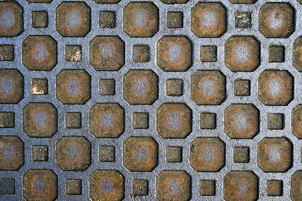 Close-up of a weathered cast iron manhole cover. The repetitive industrial texture of squares and octagons woven from bluish metal and brown rust.