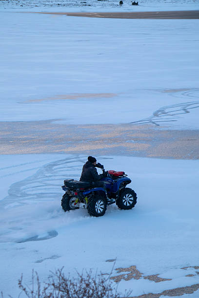 Naryn, Kyrgyzstan - December 13, 2024: A man riding a blue ATV across a snow-covered, frozen plain or lake surface, leaving tracks behind the vehicle