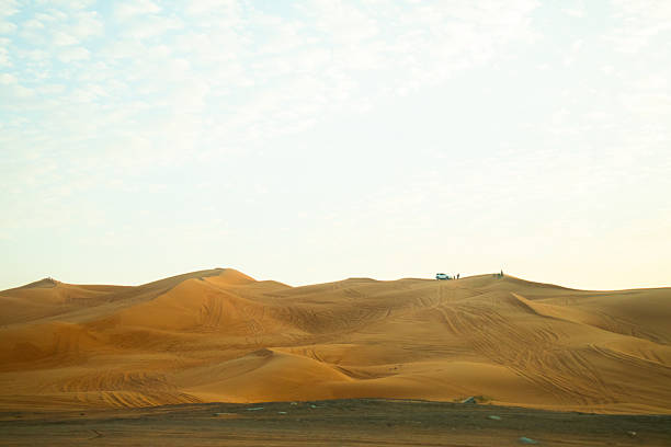 Dubai, UAE - July 13, 2024: A vast, sweeping desert landscape of massive orange sand dunes with tire tracks and distant vehicles and people visible on a ridge