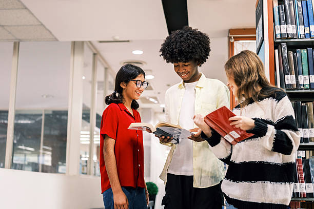 istock Three cheerful students stand in a bright library hallway, smiling and comparing books beside tall shelves, symbolizing youth education, learning resources, and academic engagement. 2246468057