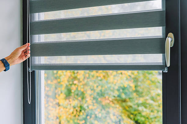 istock A woman opens black roller blinds on a window in a room. A woman's hand pulls a cord, closing the roller blinds in the room. 2246472418