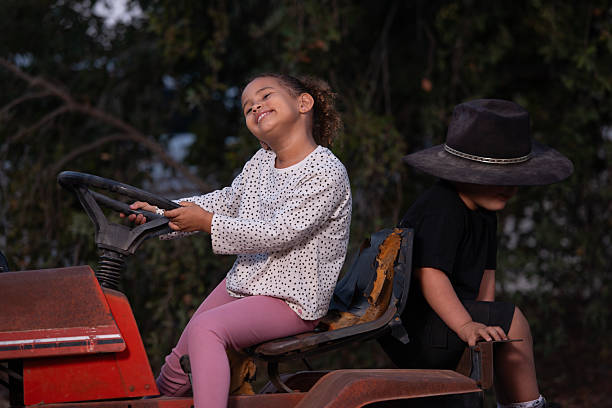 A Caucasian man in his 30s drives a tractor on his family's farm in rural Oregon while holding his elementary age daughter and preschool age son on his lap.