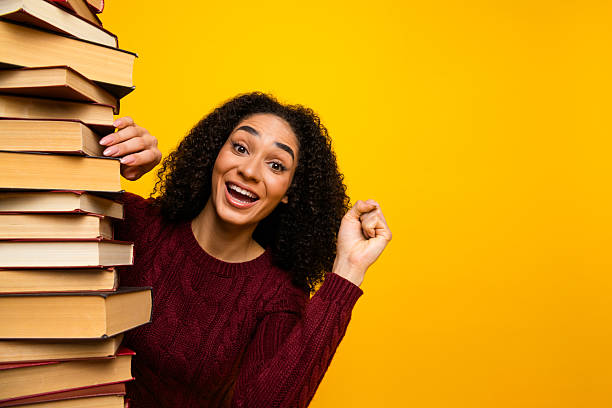 istock Young mixed race female student with joyful smile carries a tall stack of book against a bright yellow background learning study lively shopping energy 2246476482