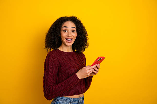 istock Cheerful young female student smiles while using a red smartphone against bright yellow background suitable for advertising fashion 2246476579