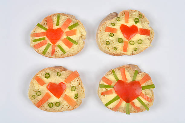 Four open-faced baguette slices are spread with butter and creatively decorated with a tomato heart, tomato slices, and sliced scallions on a white background