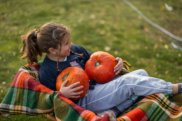 Joyful Little Girl Lying on a Plaid Blanket and Holding Pumpkins Outdoors on a Fall Afternoon