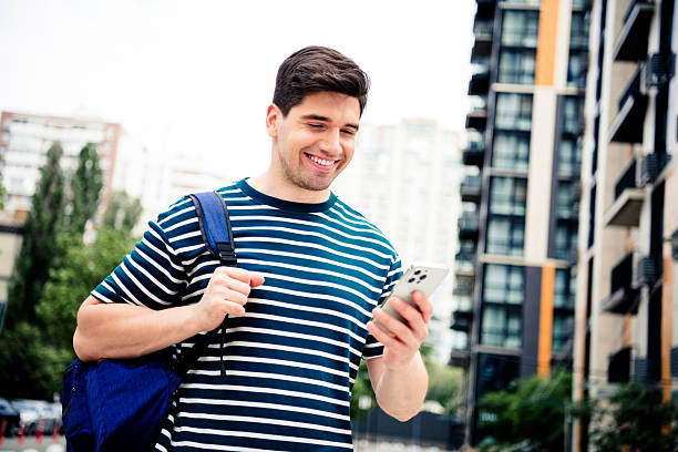 istock Young man in striped shirt walking outdoors, smiling while using smartphone in urban residential area on sunny day 2246477330