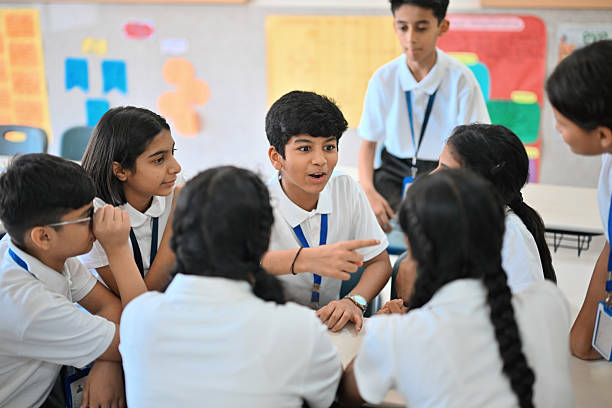 A group of middle school students gathers around a desk during recess, chatting and goofing around while one boy casually sits on the desk and gestures animatedly. The relaxed scene captures friendship, timepass, and carefree school break vibes -perfect for themes of youth, leisure, campus life, and social interaction.