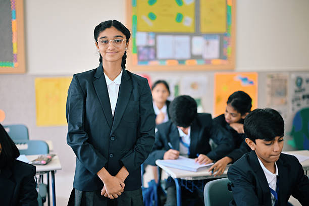 A middle school girl in a formal school blazer and glasses stands confidently in a bright classroom, smiling at the camera. Classmates work in the background, highlighting themes of education, student leadership, academic success, and back-to-school. Ideal for learning, schooling, uniforms, and youth empowerment.
