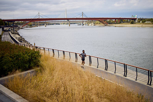 A man jogs along a riverside path with a striking bridge in the distance, urban park atmosphere.