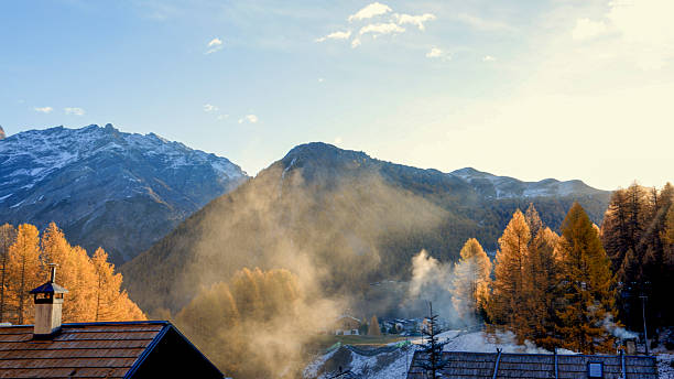 Steam rises up from houses in village at dawn, snowcapped mountains and forest in autumn, European Alps