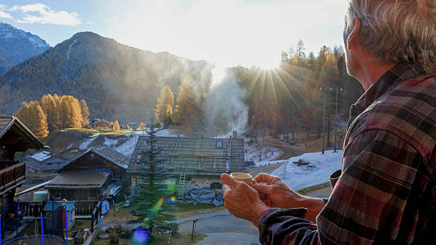 Senior man enjoys hot beverage on chalet deck at sunrise, village below and snowcapped mountains and forest in distance, autumn, European Alps