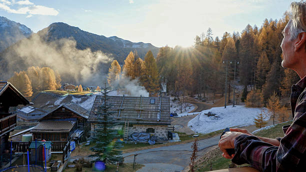 Senior man watches sunrise over village, distant mountains and larch tree forest from chalet deck, European Alps