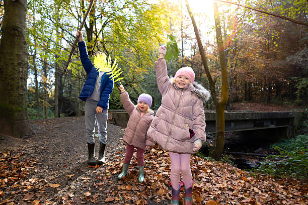 A full shot of two sisters and a brother playing energetically in a forest during a beautiful autumn day. The ground is covered in a thick carpet of fallen brown and red leaves. The children are dressed warmly in coats, hats, and boots, located in Northumberland, England.