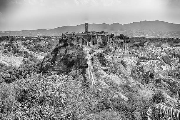 A historic village is perched atop a steep, eroded hill, connected by a long pedestrian bridge. Lush green vegetation and barren badlands surround the iconic Italian landmark under a clear sky.