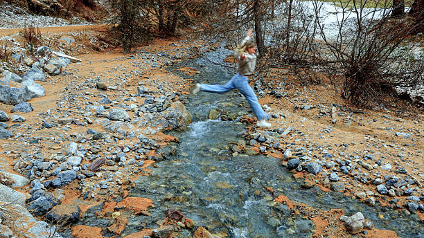 Young woman jumps over stream, autumn leaf colour in larch forest, between Livigno and St Moritz, European Alps