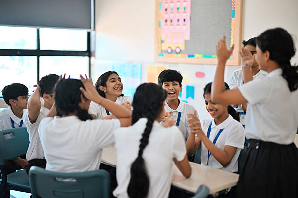 A circle of schoolchildren gathers around a table, all reaching in with their hands as if playing a fast-paced group game. One boy is sitting slightly elevated on the table, talking or calling out to the others, while the rest look up at him, smiling and engaged.